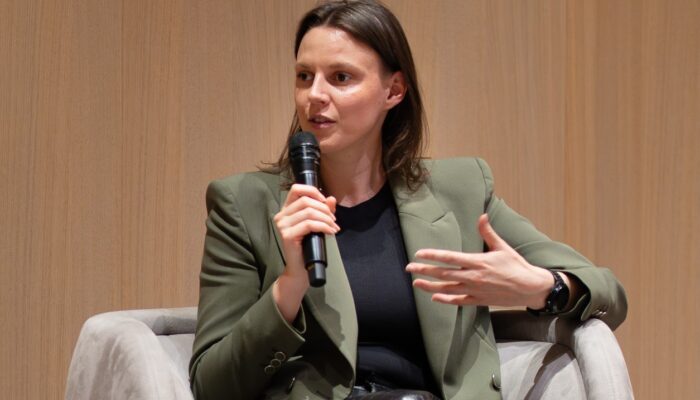 Nahaufnahme einer Frau, die auf einem Sessel sitzt und während einer Podiumsdiskussion in ein Mikrofon spricht. Sie trägt ein olivgrünes Sakko über einem dunklen Oberteil und gestikuliert mit der freien Hand vor einer hellen Holzpaneel-Wand.