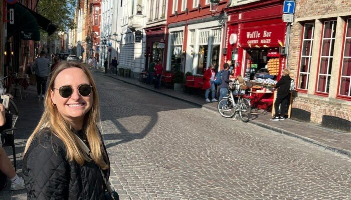 Eine lächelnde Frau steht auf einer sonnigen Kopfsteinpflasterstraße mit bunten Altstadthäusern im Hintergrund.