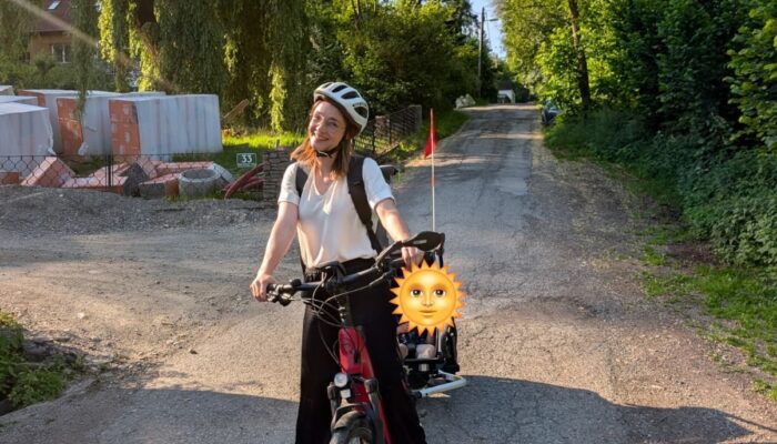 Eine lächelnde Frau mit Fahrradhelm steht mit ihrem Fahrrad auf einer ruhigen, von Bäumen gesäumten Straße. Am Fahrrad ist ein Kindersitz mit einem Kind befestigt, dessen Gesicht durch ein Sonnen-Emoji verdeckt ist. Die sommerliche Umgebung ist von üppigem Grün und warmem Sonnenlicht geprägt.
