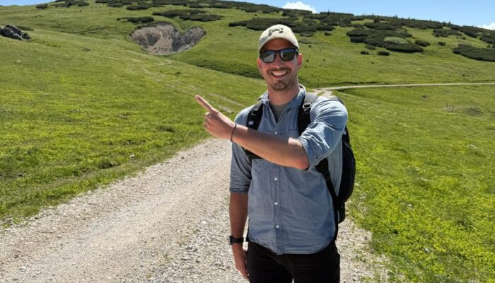 Mann mit Sonnenbrille, Rucksack und Kappe steht auf einem Schotterweg in den Bergen, lächelt und zeigt mit dem Finger auf den Hang im Hintergrund.