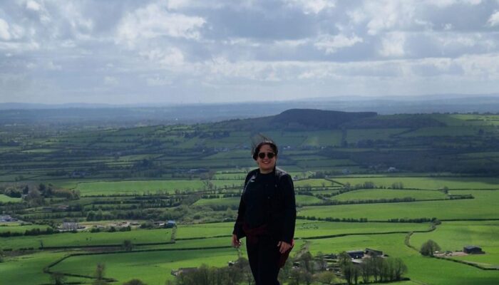 Frau auf einem Felsen mit Blick über eine weitläufige grüne Landschaft unter bewölktem Himmel.