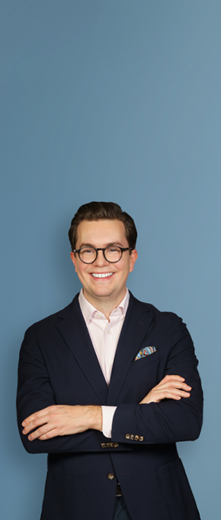 Eine lächelnde Person mit kurzen, zurückgekämmten Haaren und Brille trägt ein dunkelblaues Sakko über einem hellen Hemd. Sie steht mit verschränkten Armen vor einem hellblauen Verlaufshintergrund und blickt direkt in die Kamera.