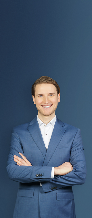 Eine lächelnde Person mit kurzen, zurückgekämmten Haaren trägt einen hellblauen Anzug über einem weißen Hemd und steht mit verschränkten Armen vor einem dunkelblauen Farbverlauf.