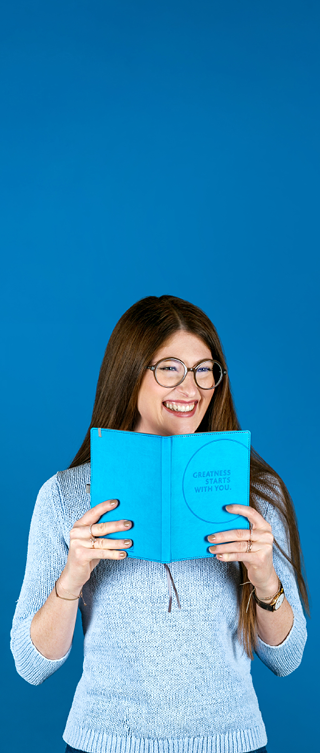 Eine lächelnde Person mit langen braunen Haaren und Brille hält ein türkisfarbenes Notizbuch vor sich und blickt darüber hinweg in die Kamera. Sie trägt einen hellblauen Pullover und steht vor einem kräftig blauen Hintergrund.