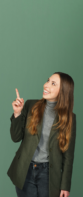 Eine lächelnde Person mit langen braunen Haaren trägt einen dunkelgrünen Blazer über einem grauen Rollkragenpullover und eine dunkle Jeans. Sie blickt nach oben links und zeigt mit dem erhobenen Zeigefinger in diese Richtung. Der Hintergrund ist ein grüner Farbverlauf.