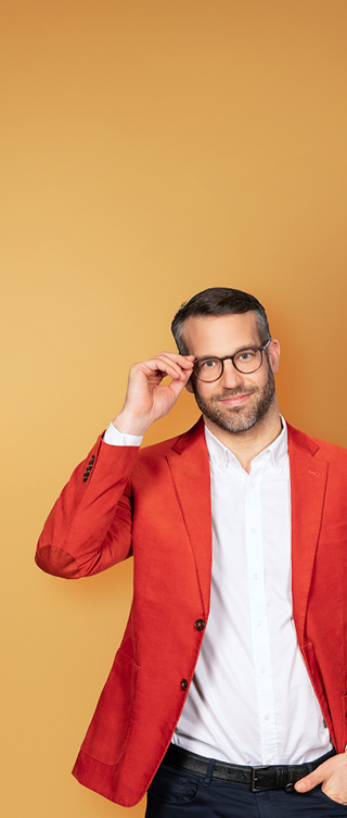 Eine lächelnde Person mit kurzen dunklen Haaren, Bart und Brille trägt ein rotes Sakko über einem weißen Hemd. Mit einer Hand fasst sie an die Brille und blickt direkt in die Kamera. Der Hintergrund ist ein goldgelber Farbverlauf.