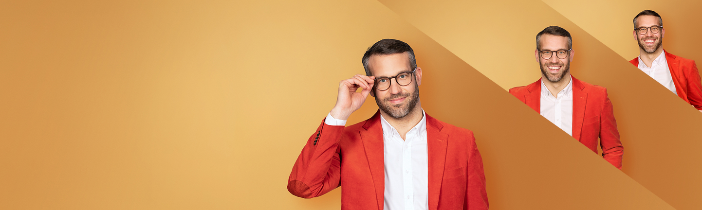 Banner mit goldgelbem Verlaufshintergrund. Im Vordergrund steht eine lächelnde Person mit kurzen dunklen Haaren, Bart und Brille. Sie trägt ein rotes Sakko über einem weißen Hemd und fasst mit einer Hand an die Brille. Rechts oben sind zwei kleinere Ausschnitte derselben Person als gestalterische Elemente eingebunden.