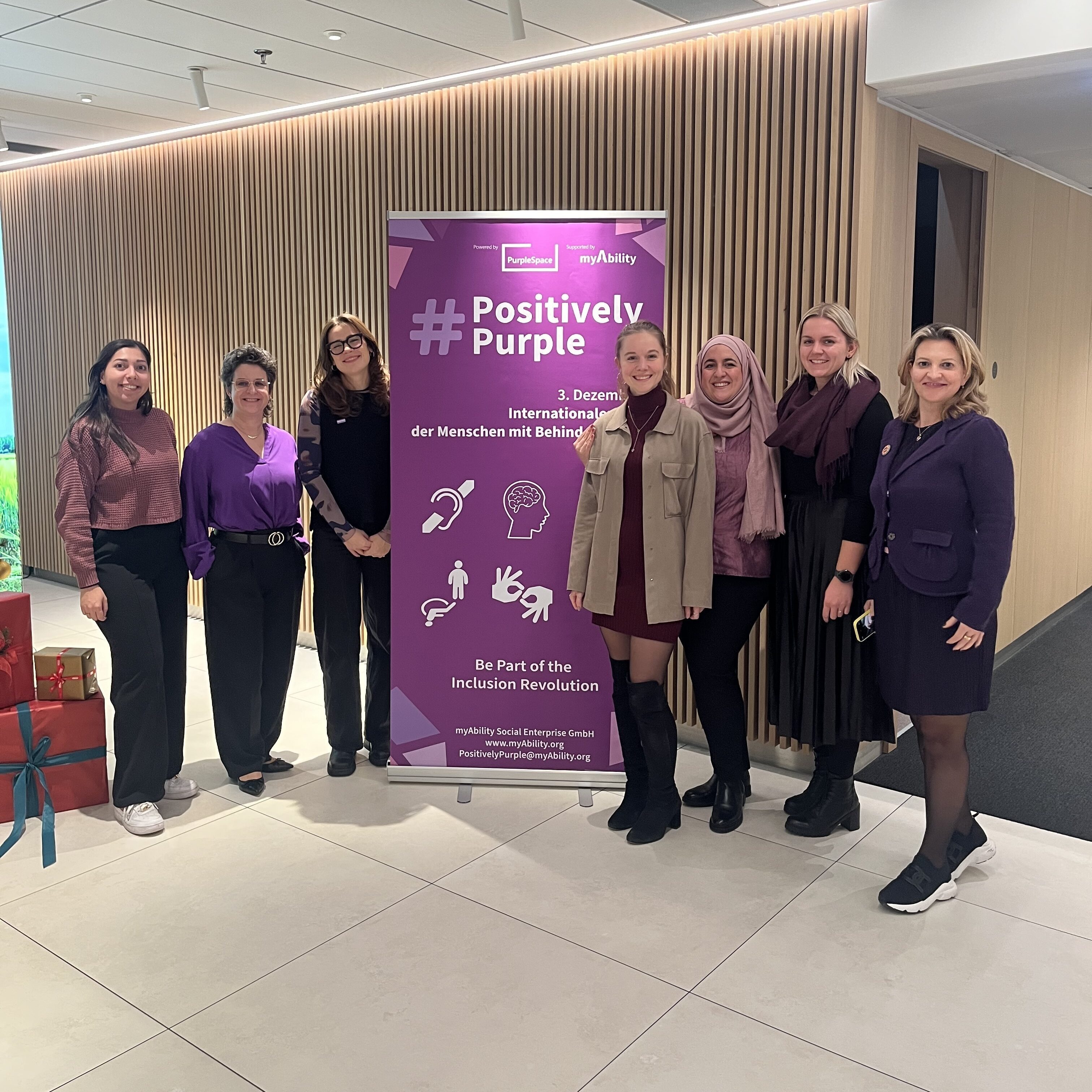 Sieben Personen posieren gemeinsam neben einem Banner der Kampagne „#PositivelyPurple" anlässlich des Internationalen Tags der Menschen mit Behinderung.