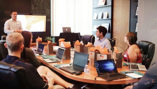 Eine Gruppe Menschen sitzt mit Laptops in einem Besprechungszimmer. Ein Mann steht vor einem Bildschirm und präsentiert etwas.