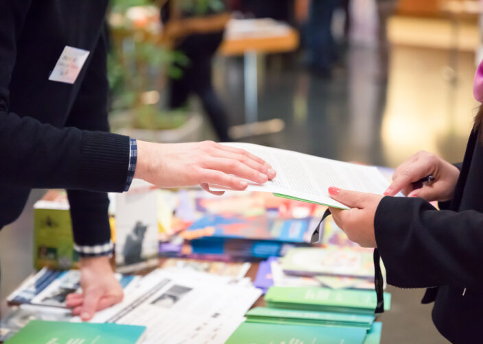 Nahaufnahme: Zwei Personen tauschen an einem Stand mit Broschüren und Flyern Informationsmaterial aus.