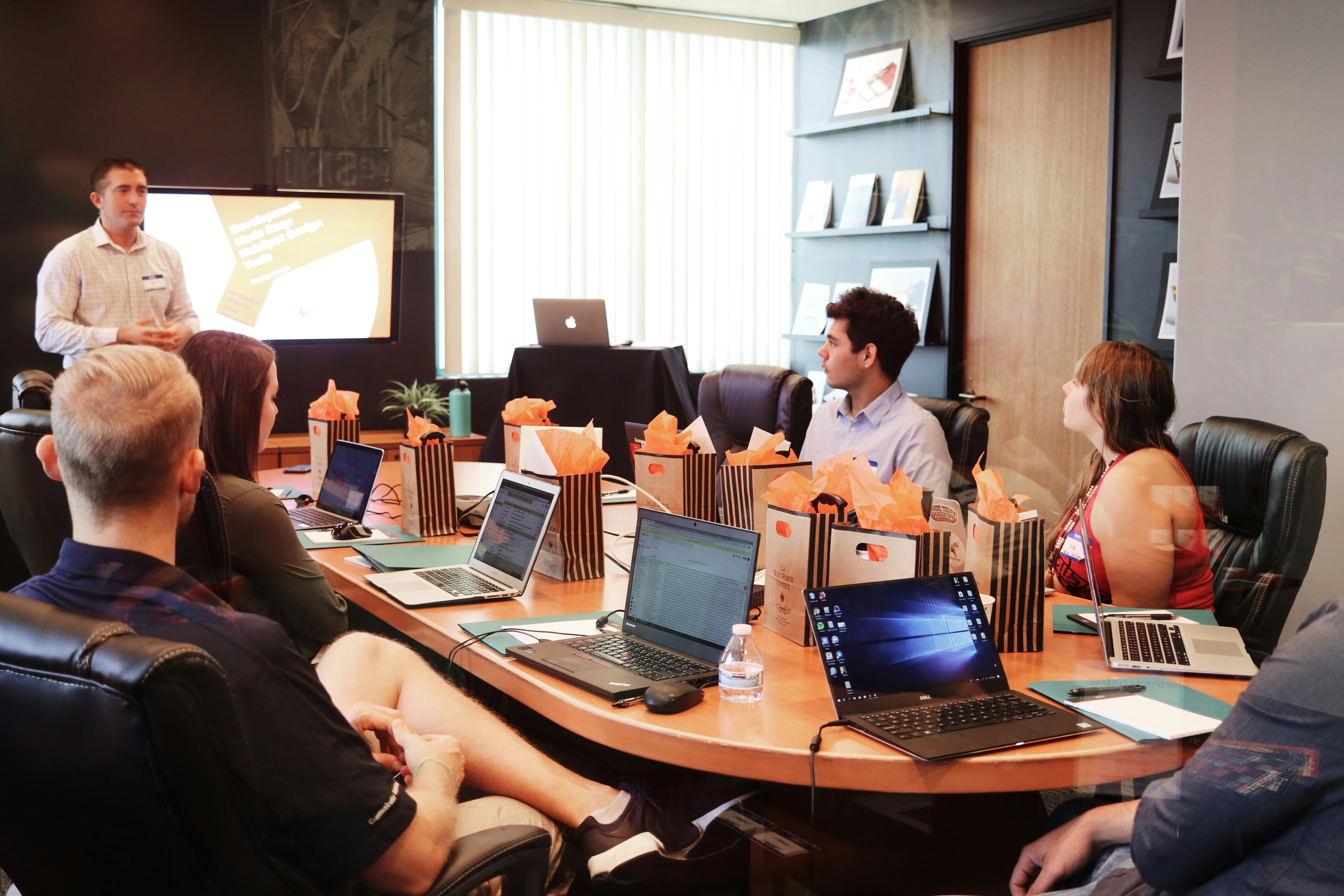 Eine Gruppe Menschen sitzt mit Laptops in einem Besprechungszimmer. Ein Mann steht vor einem Bildschirm und präsentiert etwas.