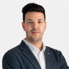 Porträt einer Person mit dunklem Haar, grauem Sakko und weißem Hemd vor hellem Hintergrund.
