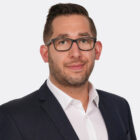 Porträt einer Person mit kurzem dunklem Haar, schwarzer Brille und leichtem Lächeln. Die Person trägt ein weißes Hemd und ein dunkelblaues Sakko und blickt direkt in die Kamera. Der Hintergrund ist hell und neutral.