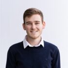 Porträt einer lächelnden Person mit kurzen, braunen Haaren, dunkelblauem Pullover und weißem Hemd vor hellem Hintergrund.