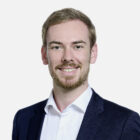 Porträt einer lächelnden Person mit kurzen, hellbraunen Haaren und einem gepflegten Bart. Sie trägt ein weißes Hemd und ein dunkelblaues Sakko und blickt direkt in die Kamera. Der Hintergrund ist hell und neutral.