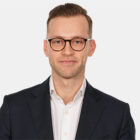 Porträt eines Mitarbeiters im dunklen Anzug mit weißem Hemd und Brille vor hellem Hintergrund