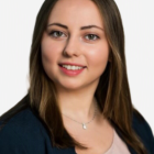 Porträt einer lächelnden Person mit schulterlangem, braunem Haar, dunklem Blazer und hellem Oberteil vor hellem Hintergrund.