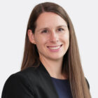 Porträt einer lächelnden Person mit langen, glatten, braunen Haaren. Sie trägt einen schwarzen Blazer über einem dunkelblauen Oberteil und blickt direkt in die Kamera. Der Hintergrund ist hell und neutral.