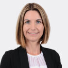 Porträt einer lächelnden Person mit schulterlangem, hellbraunem Haar, schwarzem Blazer und weißer Bluse vor hellem Hintergrund.
