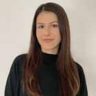 Porträt einer Person mit langen, dunklen Haaren und schwarzem Oberteil vor hellem Hintergrund.