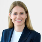 Porträt einer lächelnden Person mit langen, glatten, blonden Haaren. Sie trägt einen dunkelblauen Blazer über einem weißen Oberteil und blickt direkt in die Kamera. Der Hintergrund ist hell und neutral.