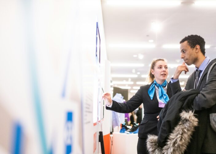 Zwei Personen im Gespräch an einem Stand: Eine Frau zeigt auf eine Informationstafel, während ihr Gegenüber aufmerksam zuhört.