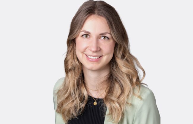 Porträt einer lächelnden Person mit hellgrünem Blazer und schwarzem Oberteil vor einem hellgrauen Hintergrund.
