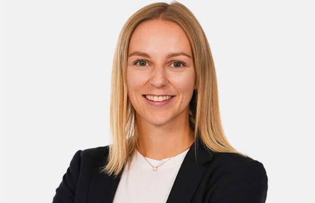 Eine junge Frau mit glattem, blondem Haar lächelt in die Kamera. Sie trägt ein dunkles Jackett über einem weißen Oberteil vor neutralem Hintergrund.