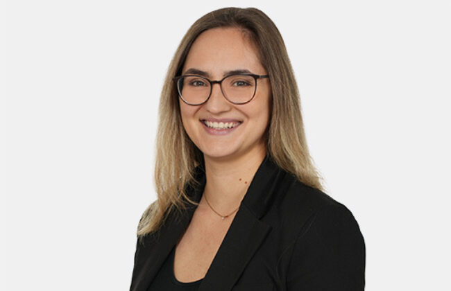 Porträt einer lächelnden Person mit schulterlangen blonden Haaren und Brille. Sie trägt ein schwarzes Oberteil und einen schwarzen Blazer und blickt direkt in die Kamera. Der Hintergrund ist hell und neutral.