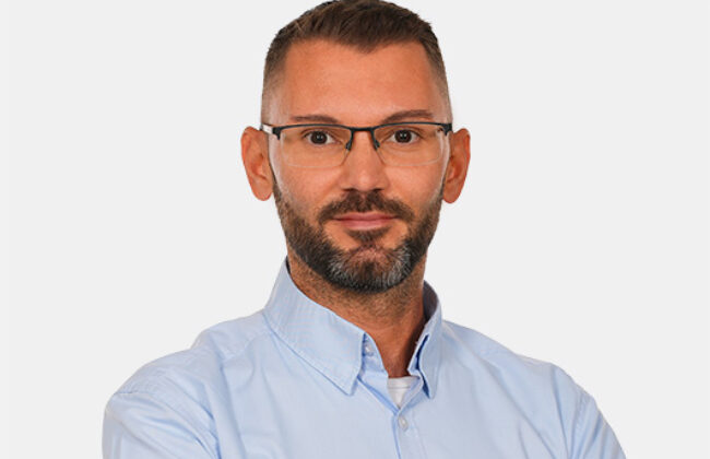 Ein Mann mit Brille und Bart trägt ein hellblaues Hemd und blickt freundlich in die Kamera, vor neutralem Hintergrund.