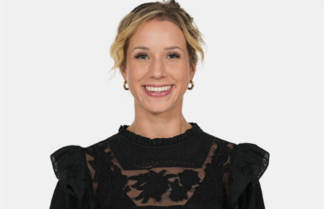 Porträt einer lächelnden blonden Frau mit hochgesteckten Haaren, die eine schwarze Bluse mit Spitzendetails trägt und vor einem hellen Hintergrund in die Kamera blickt.